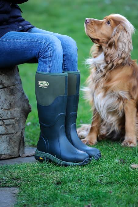 A person wearing dark green and navy rubber rain boots sits on a stone outdoors next to a golden retriever dog on a green lawn. The boots feature a green upper section with a visible 'WELLINGTON' brand logo.